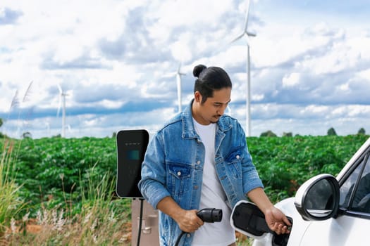 Progressive man with his electric car, EV car recharging energy from charging station on green field with wind turbine as concept of future sustainable energy. Electric vehicle with energy generator.