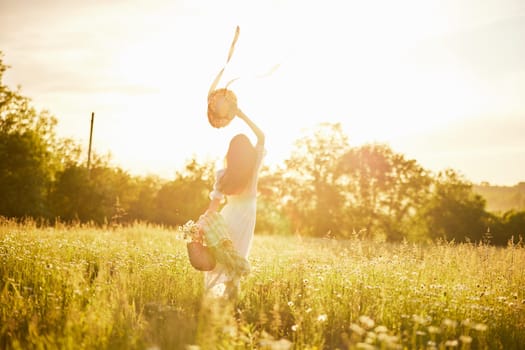 a woman in a light dress stands in a field in the warm rays of the setting sun holding a hat over her head. High quality photo