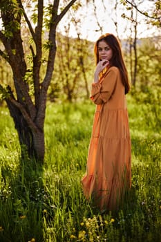 a beautiful woman stands in a long orange dress, in the countryside, near a flowering tree, during sunset, and holds her hand near her face. Vertical photography. High quality photo