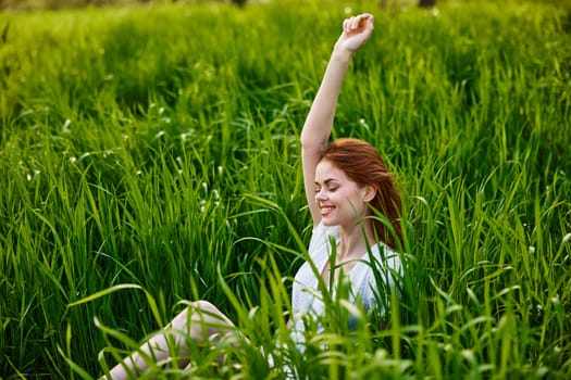 a cute woman in the summer high grass sits in a light dress happily raising her hand up. High quality photo