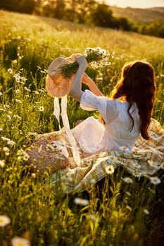 a woman sitting with her back to the camera in nature in a light dress holds a hat and a bouquet of daisies in her hands. High quality photo