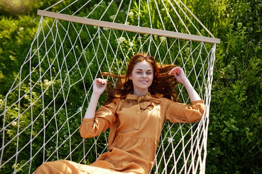 a beautiful, elegant woman lies in a long orange dress on a mesh hammock resting in nature, illuminated by the warm sunset light, smiling happily. Horizontal photo taken from above. High quality photo