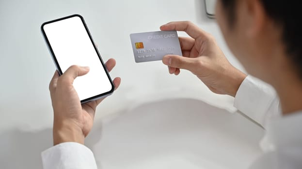 Over shoulder view of man holding credit card and smartphone, shopping online or doing online banking transaction.