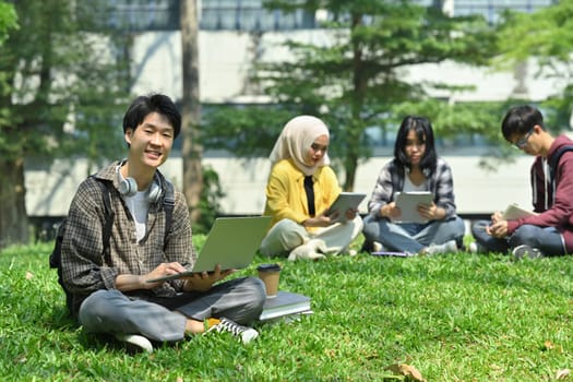 Young asian man student using laptop on green grass in front of university building. Education, technology and lifestyle concept.