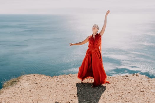 Side view a Young beautiful sensual woman in a red long dress posing on a rock high above the sea during sunrise. Girl on the nature on blue sky background. Fashion photo.