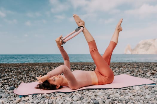 Middle aged well looking woman with black hair doing Pilates with the ring on the yoga mat near the sea on the pebble beach. Female fitness yoga concept. Healthy lifestyle, harmony and meditation.