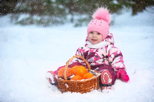 A little girl in winter clothes is sitting on the snow with a basket of oranges.
