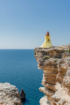 Woman in a yellow dress on the sea. Side view Young beautiful sensual woman in yellow long dress posing on a rock high above the sea at sunset. Girl in nature against the blue sky.