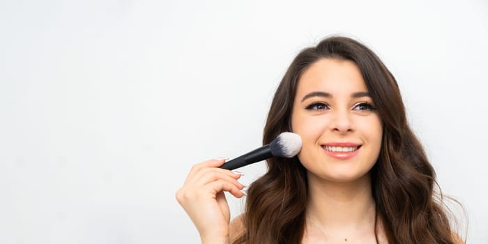 Banner with smiling young woman holding a cosmetic brush near her face. Makeup artist applying face powder.