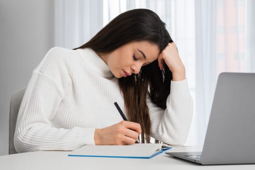 Tired student makes notes in the textbook, prepares for an exam or does homework sitting at a desktop at home.