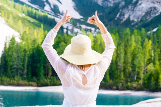 Woman in the straw hat and white dress shows the sign Like on the background of mountains, forest and lake.