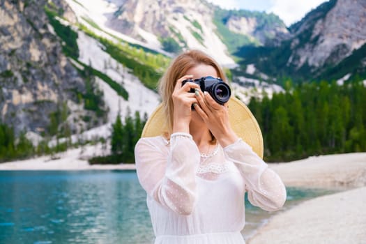 Portrait of a blonde hair female photographer covering her face with the camera on the background of Lake Braies and the Dolomites