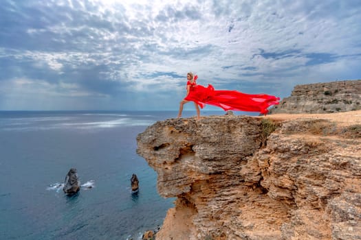 A woman in a red silk dress stands by the ocean, with mountains in the background, as her dress sways in the breeze