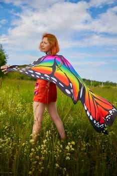 Adult girl with red hair and butterfly wings having fun and joy in meadow or field with grass, flowers on sunny summer day
