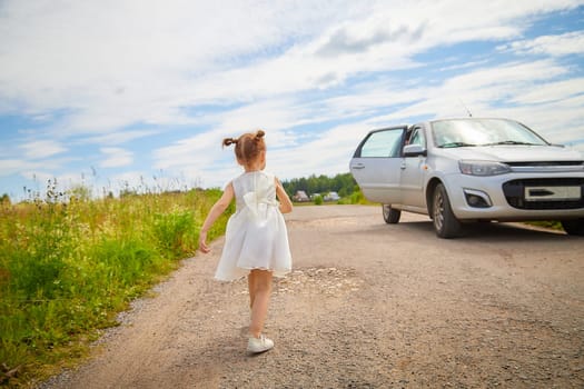 Portrait of little girl with Asian eyes on a road near car in a meadow or field with grass and flowers on sunny summer day. Rural, rustic journey