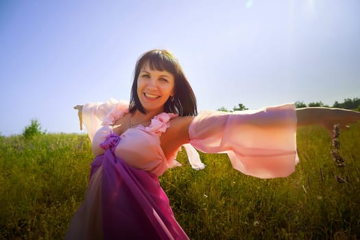Beautiful girl in lush pink ball gown in green field during blooming of flowers and blue sky on background. Model posing on nature landscape as princess from fary tale