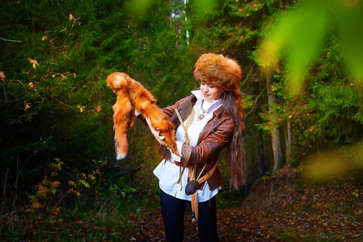 Girl in a leather jacket, a big red fox fur hat and with the skin of a fox killed on the hunt in the forest in autumn. A female model poses as fabulous royal huntress on nature hunt at photo shoot