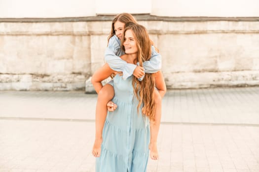 Mother of the daughter walks playing. Mother holds the girl on her back, holding her legs, and her daughter hugs her by the shoulders. Dressed in blue dresses