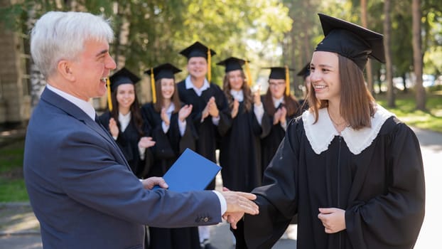 The teacher shakes hands with the student and presents the diploma outdoors. A group of university graduates