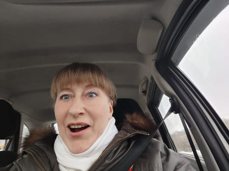 Portrait of adult cute funny blonde woman in warm clothes taking a selfie inside the car and driving it. The concept of pleasant trip in spring, autumn, winter