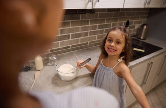 Adorable beautiful little girl smiling with cheerful toothy smile looking at her mom while mixing ingredients with flour in glass bowl with a whisk at home kitchen, preparing dough for Shrove pancakes