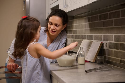 Adorable happy mother smiles to her cute little daughter mixing the ingredients in a bowl while preparing pancake dough. Motherhood, mom teaching her daughter to cook pancakes for Shrove Tuesday