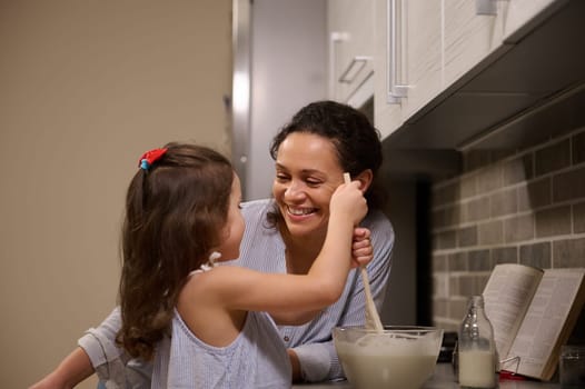 Adorable little girl having fun, mixing ingredients and kneading dough in a glass bowl while her loving happy mother teaching her to cook pancake dough.