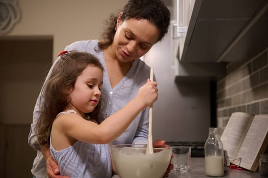 Adorable little girl having fun, concentrated on mixing ingredients and kneading dough in a glass bowl while her loving happy mother teaching her to cook pancake dough. Shrove Tuesday concept