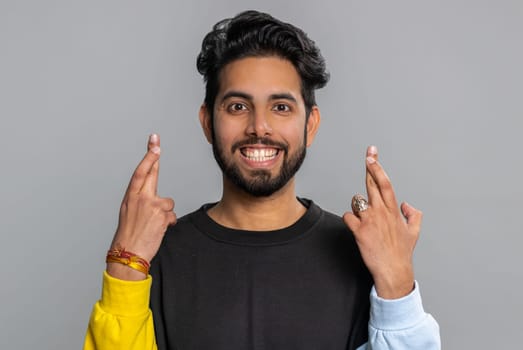 Indian young man crossed fingers looking at camera asking for good lucky news, wishing good exam results, dreaming about win victory in lottery jackpot. Hindu guy isolated on gray studio background