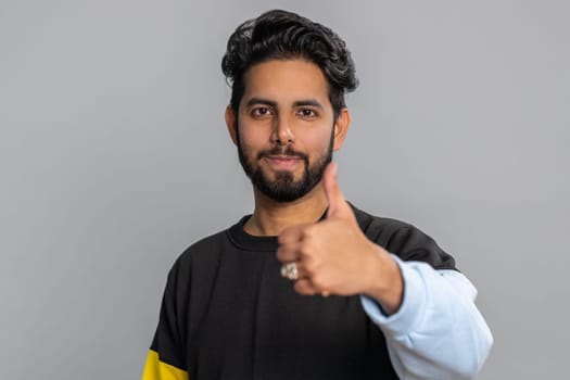 Like. Indian young man in sweater raises thumbs up agrees with something, gives positive reply recommends advertisement likes good. Handsome bearded hindu guy isolated alone on gray studio background