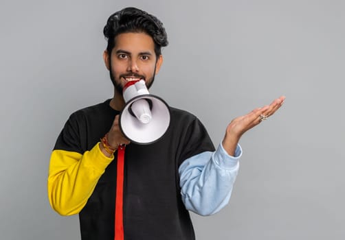 Indian man talking with megaphone, proclaiming news, loudly announcing advertisement, warning using loudspeaker to shout speech, pointing empty place. Copy-space. Handsome guy on gray wall background
