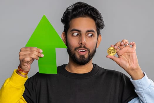 Indian man winner holding arrow sign pointing up showing golden bitcoins, cryptocurrency investment, mining BTC money exchange increase. Hindu guy showing up indicating successful development progress
