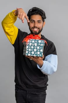 Positive smiling young indian man presenting birthday gift box stretches out hands, offer wrapped present career bonus, celebrating party, sale. Bearded hindu guy isolated on gray studio background