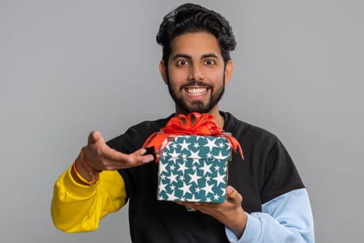 Positive smiling young indian man presenting birthday gift box stretches out hands, offer wrapped present career bonus, celebrating party, sale. Bearded hindu guy isolated on gray studio background