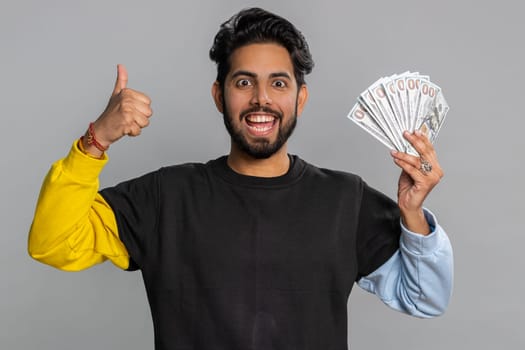 Rich pleased boss hindu man waving money dollar cash banknotes bills like a fan, success business career, lottery winner, big income, wealth. Handsome indian guy isolated on gray background, indoor