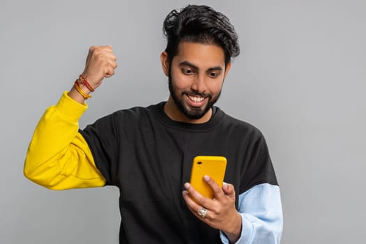 Happy excited joyful indian man in black sweater use smartphone typing browsing shouting say wow yes found out great big win, good news, lottery goal achievemen, celebrating success, winning game