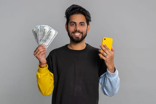 Indian man looking smartphone display sincerely rejoicing win, receiving money dollar cash banknotes, success lottery jackpot luck, income salary wage. Hindu guy isolated on studio gray background