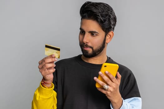 Sincere indian man customer using credit bank card and smartphone while transferring money, purchases online shopping, payment. Finance and internet. Handsome guy isolated alone on gray background