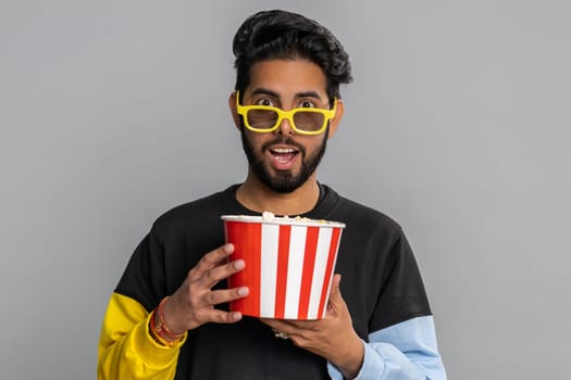 Excited indian man in 3D glasses eating popcorn and watching interesting tv serial, sport game, film, online social media movie content. Hindu guy enjoying domestic entertainment on gray background