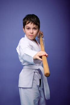 Caucasian child boy in a white kimono fights with a wooden weapon in aikido training on a violet background with copy space for advertising text. Healthy lifestyle and martial arts concept