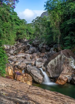 A remote and indomitable terrain surrounds a channel of flowing water, where a majestic river of authentic large round stones smoothed over time by history descends from the mountain, surrounded by lush green jungle and featuring silky smooth water in a long exposure photo.