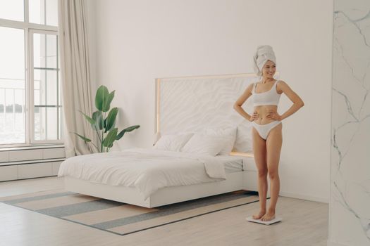 Shot of healthy slim young woman in white underwear with head wrapped in bath towel standing on scales in bedroom, happy to see her weight loss. Healthy lifestyle and body care concept