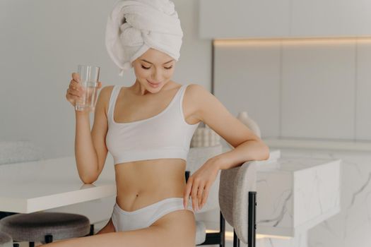 Fit attractive young woman sitting in kitchen while drinking glass of water, rehydrating herself after shower routine procedure, wears underwear and towel wrapped on head. Healthy morning habbits
