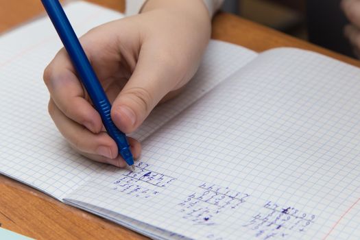 The girls hands with a blue fountain pen solve examples in close-up. The student completes the task in teradi. A schoolboy teaches lessons at a school desk. Warm soft daylight.