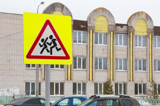 Road sign means be careful children. Against the background of the school building and cars standing nearby. Humans run on a white background in a red triangle and a yellow square. Warning sign.