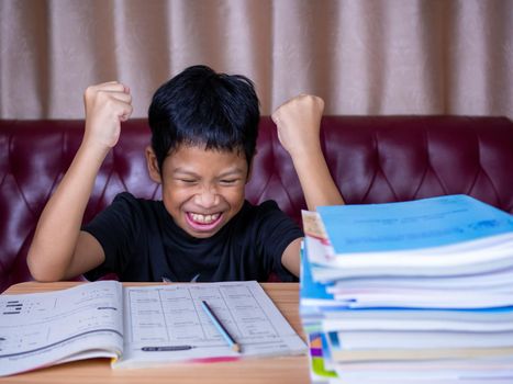 The boy was very happy to finish his homework. He sat on a wooden table and had a stack of books next to it. The background is a red sofa and cream curtains.