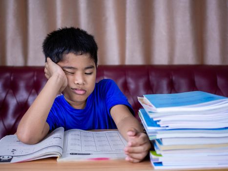 A boy is tired of reading a book on a wooden table. with a pile of books beside The background is a red sofa and cream curtains.