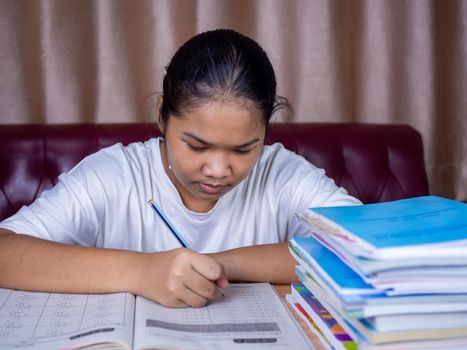 girl doing homework on a wooden table and there was a pile of books next to it The background is a red sofa and cream curtains.