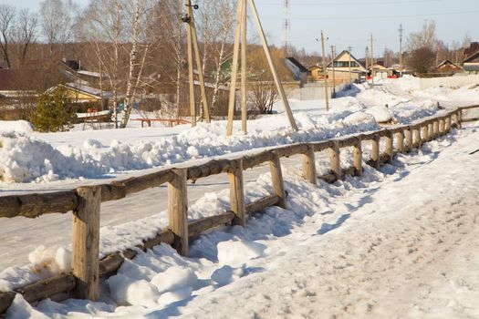 Beautiful countryside in winter with wooden fence and houses in the distance. Wonderful rural landscape with wooden houses and a snow-covered road behind a hedge.