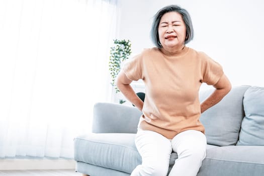 An agonizing senior woman experiencing back pain while sitting on her sofa at home and holding her back.